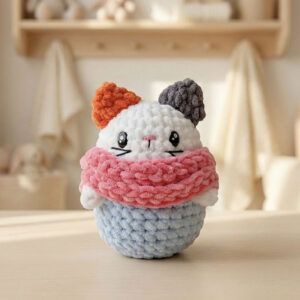 Gatita amigurumi Michi tejida con hilo suave, con bufanda rosa y base azul, sobre fondo cálido de madera.